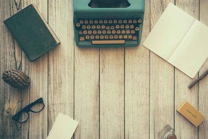 Photo of an old type writer, notebooks, glasses, a pen, all on a wooden table.