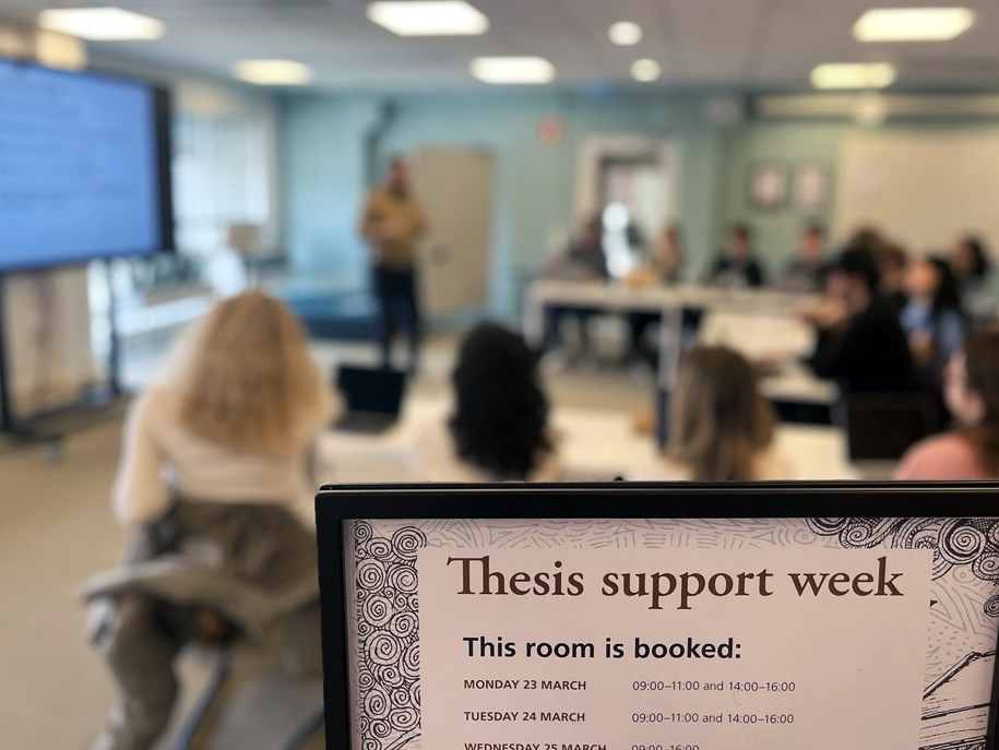 A framed poster reading: Thesis support week, and a blurry background with people in a class room